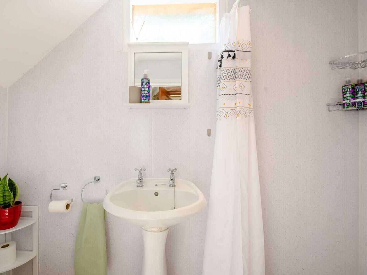 A bathroom with a pedestal sink, a shower curtain, a towel rack, a toilet paper holder, and a shelf with toiletries.