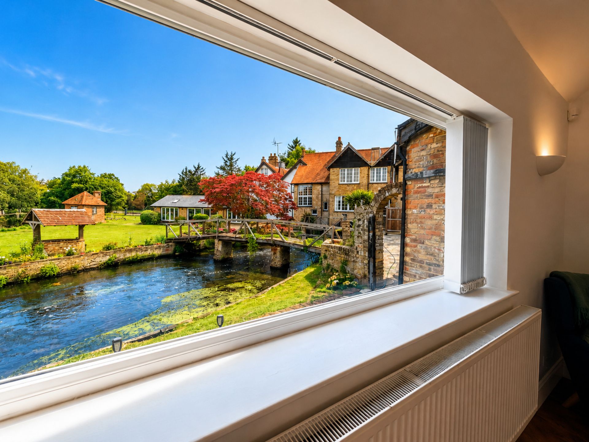 Blick aus einem Fenster auf einen Fluss mit einer Brücke, einen Baum mit roten Blättern und traditionelle Gebäude.