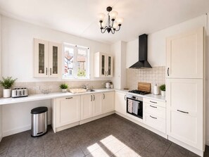 A modern kitchen with white cabinetry, a stainless steel trash bin, a gas stove, and a window providing natural light. - Luxury 5BDR Riverside Retreat near Uxbridge (Harefield)