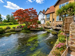 Ein Haus am Flussufer mit einer Stegenbrücke, einem Garten mit blühenden Pflanzen und einem traditionellen Gebäude mit vielen Fenstern.