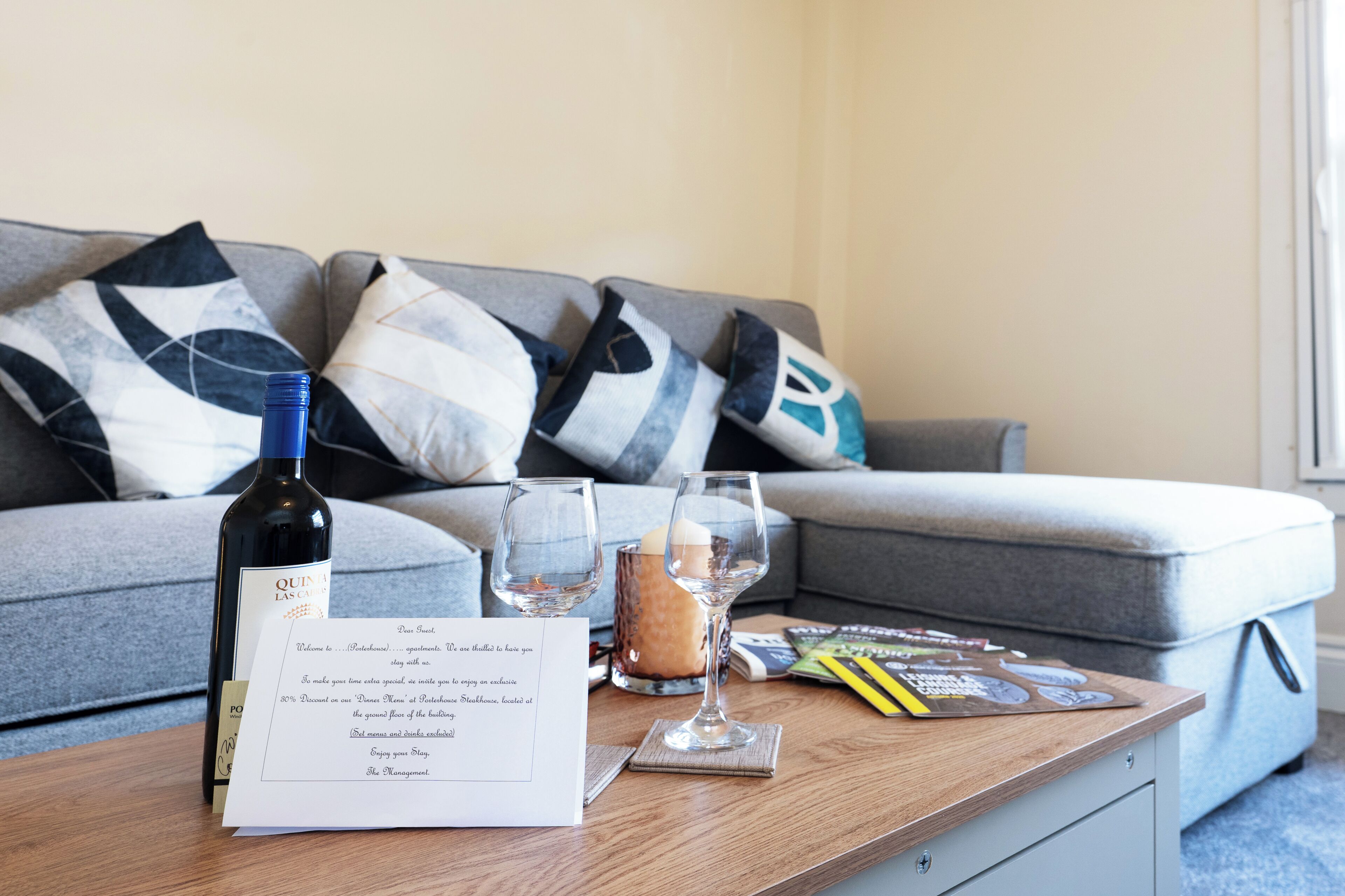 A room with a wooden coffee table, a bottle of wine, two wine glasses, a candle, and a welcome message.