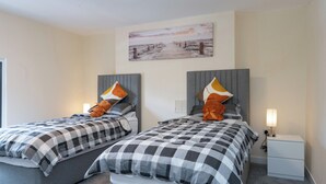 A hotel room with two beds, each with a grey headboard and a checkered bedspread. There is a painting on the wall above the beds, and a nightstand with a lamp on the right side. - The Porterhouse - Residence II (Winchester)