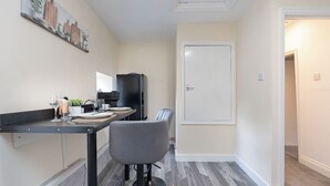 A compact kitchen with a dining area, featuring a refrigerator, a dining table with chairs, and a wall-mounted shelf with decorative items. - The Porterhouse - Residence II (Winchester)
