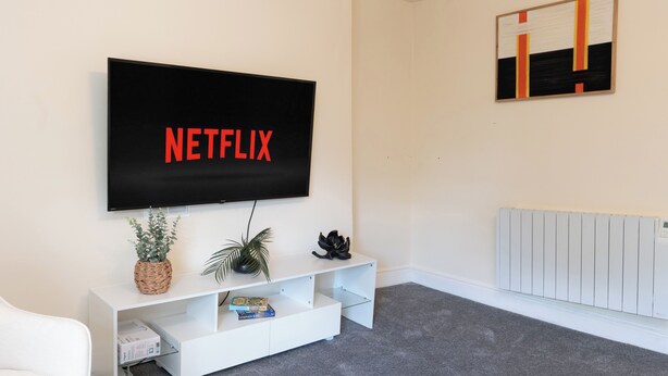A living room with a flat-screen TV mounted on the wall displaying 'NETFLIX' in red letters, a white entertainment unit with a plant and decorative items, and a radiator on the wall. - The Porterhouse - Residence II (Winchester)
