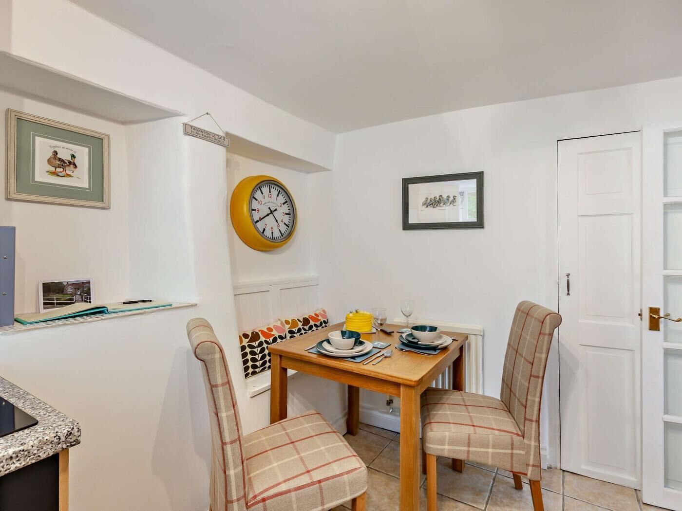 A dining area with a wooden table, two plaid chairs, a yellow clock, and framed artwork on the walls.