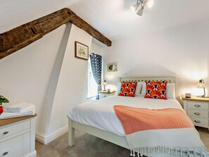Ein Schlafzimmer mit schrägen Wänden, einem Bett mit gemusterten Kissen, einem Nachttisch mit Lampe und einem Fenster mit Vorhängen.