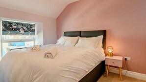 Ein Schlafzimmer mit einem Bett, Kissen, einem Nachttisch mit Lampe, einem Fenster mit Kunstwerk und einem Teppichboden.