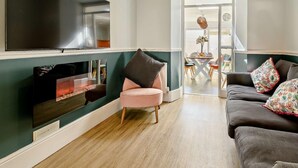 Ein Wohnzimmer mit Flachbildfernseher, einem rosafarbenen Sessel, einem dunklen Sofa und einem Essbereich im Hintergrund.