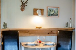 A dining area with a wooden table set for two, a lit wall lamp, and a rustic decor including a deer head and a framed painting. - 1 Bed in Hexham (oc-89085) (Bellingham)