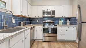A modern kitchen with white cabinets, a stainless steel microwave, oven, and refrigerator, a granite countertop, and a blue-tiled backsplash.