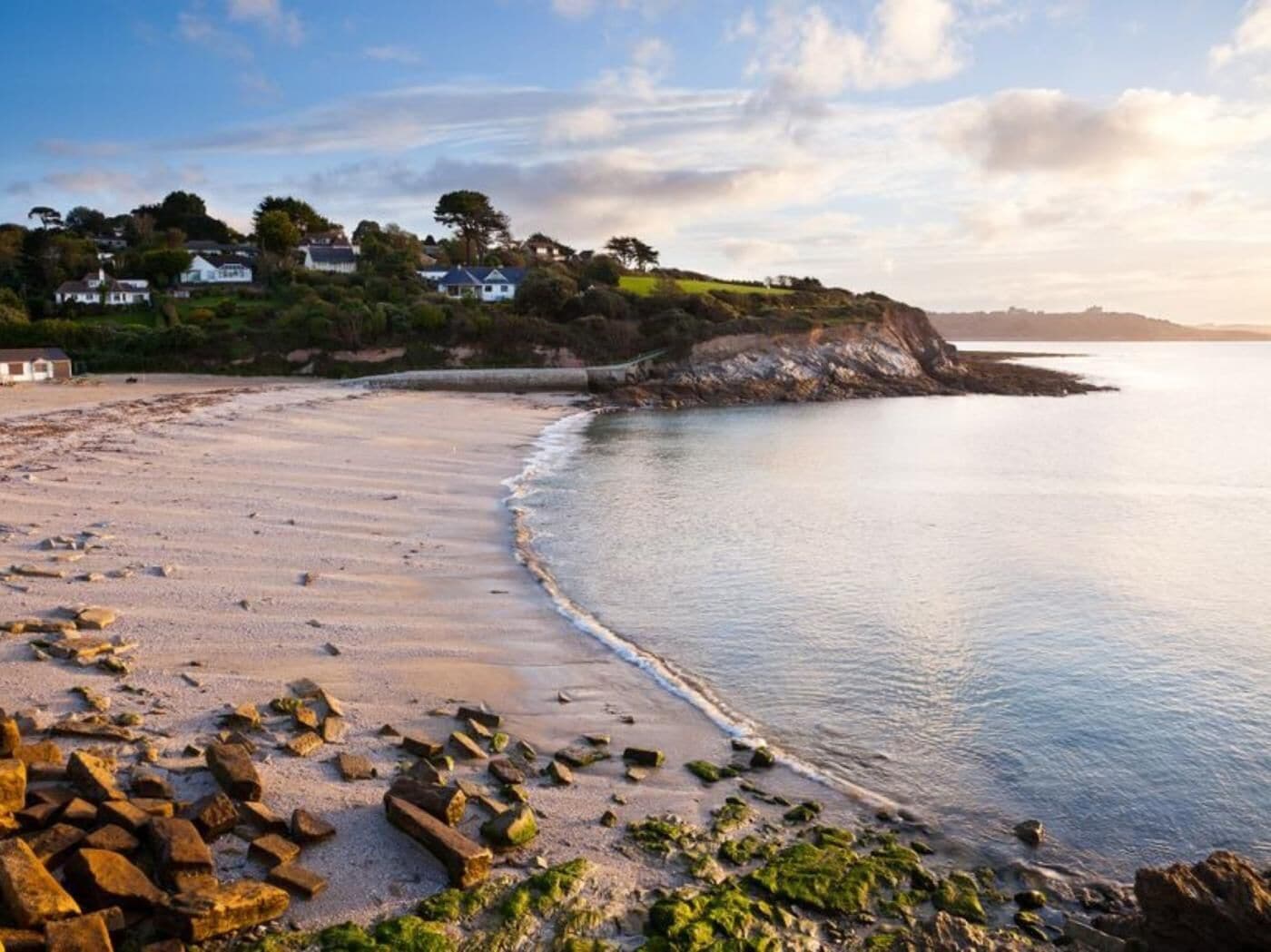 Ein Küstenstrand mit verstreuten Felsen, einem ruhigen Meer und Häusern an einem Hang.