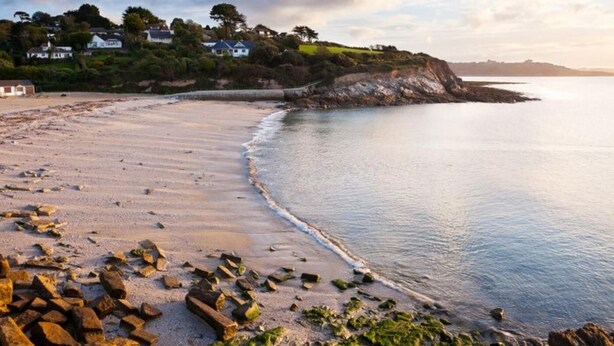 Uma praia costeira com pedras espalhadas, mar calmo e casas em uma encosta.