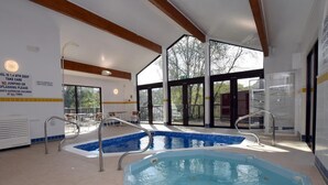 Piscina interior com paredes de vidro transparente, uma banheira de hidromassagem separada e uma área ao lado da piscina com mesas e cadeiras.