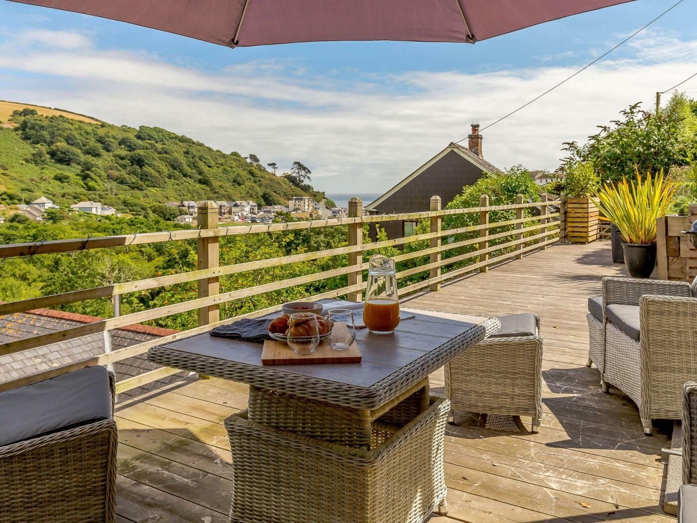 Um deck de madeira com móveis de vime, uma mesa com um jarro de vidro e vista para uma encosta.