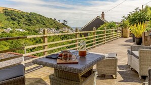A wooden deck with wicker furniture, a table with a glass pitcher, and a view of a hillside. - 4 Bed in Torpoint (oc-89135) (Deviock)