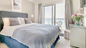 A bedroom with a bed, a bedside table with a vase of flowers, a chair, and a view of the sea through a large window. - 1 Bed in Newquay (oc-89230) (Newquay)