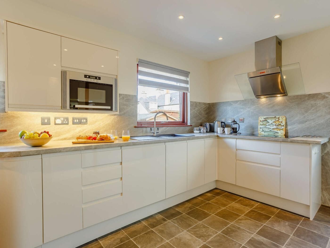 A modern kitchen with white cabinetry, a stainless steel range hood, a microwave, and a window with blinds.