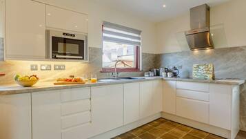 A modern kitchen with white cabinetry, a stainless steel range hood, a microwave, and a window with blinds.