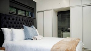 Ein Schlafzimmer mit einer gesteppten Kopfteil, weißer Bettwäsche, einer braunen Tagesdecke und einer Schiebetür zum Kleiderschrank.