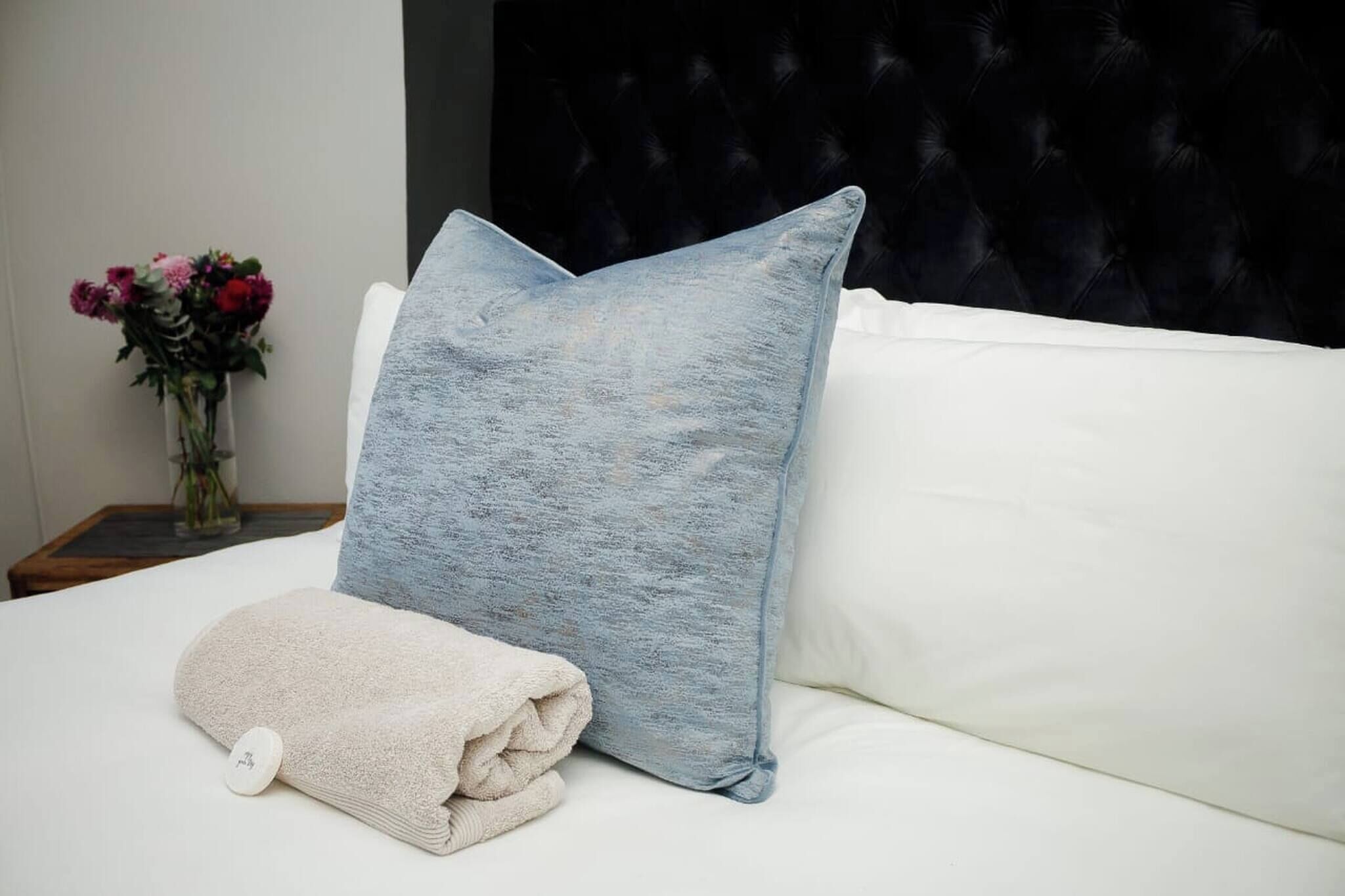 Een bed met een wit kussen en een blauw kussen, een opgevouwen witte handdoek en een vaas met bloemen op een nachtkastje.