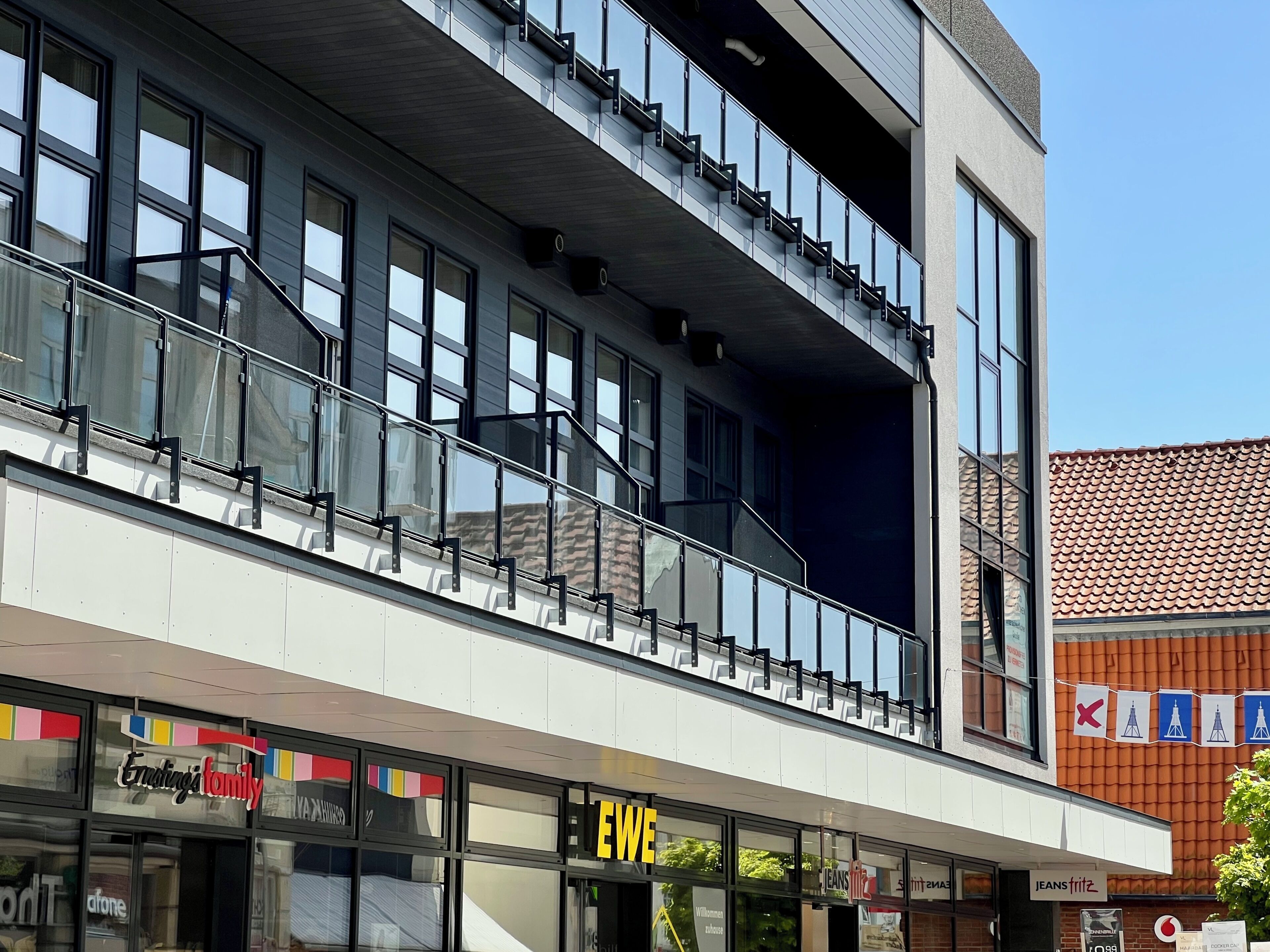 A modern building with a glass facade, a balcony, and storefronts including 'EWE' and 'Jeans Fritz'.