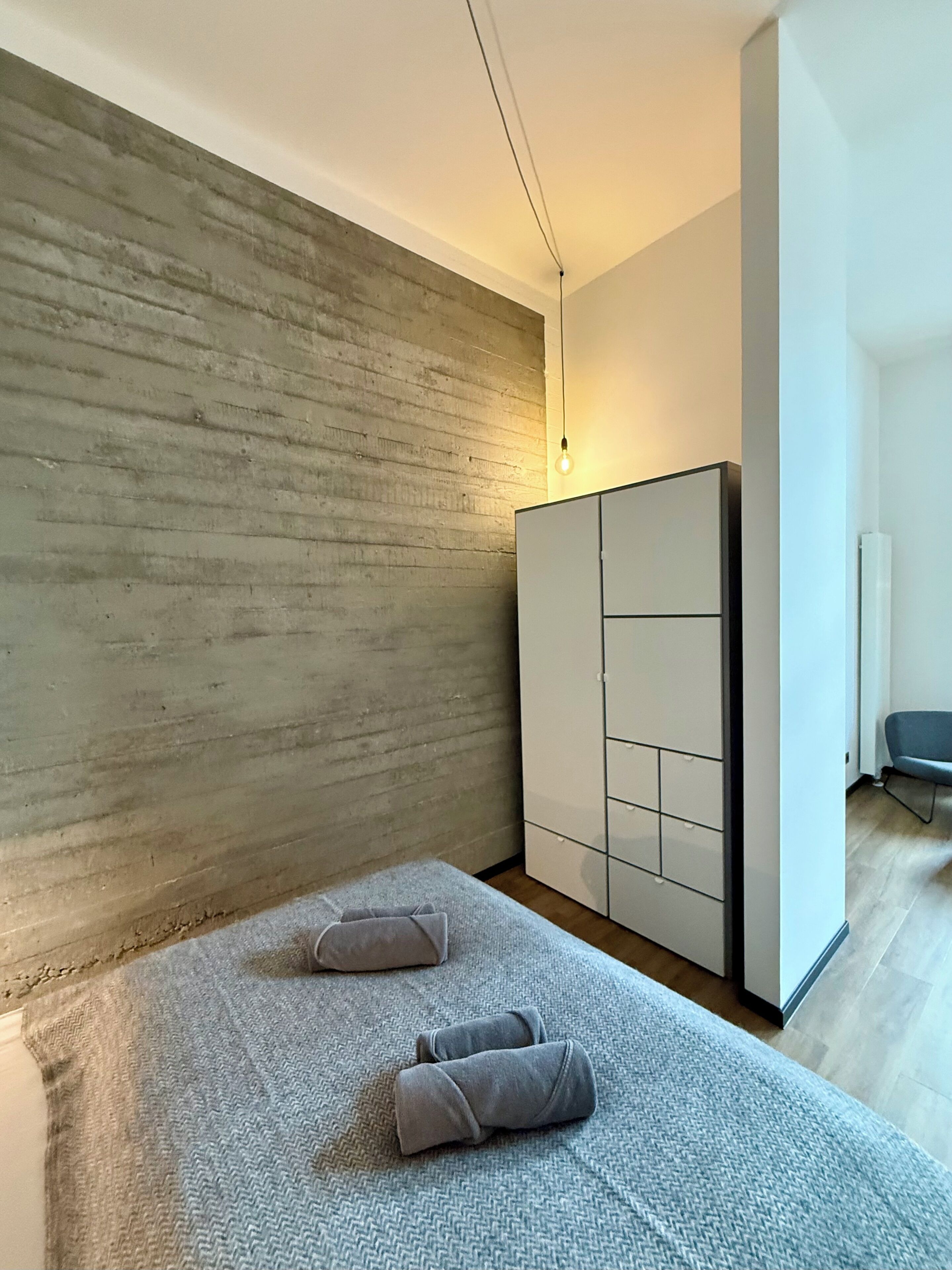 Ein modernes Schlafzimmer mit einer Betonwand, einem Bett mit grauer Bettwäsche, einem Kleiderschrank und einem Sessel.
