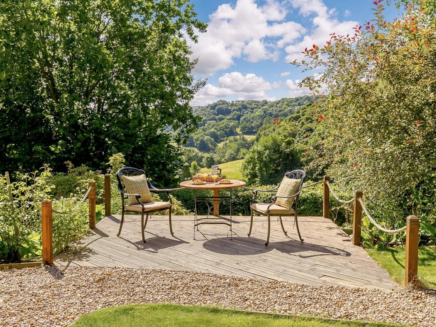 A wooden deck with two chairs and a table, surrounded by greenery and a scenic view.