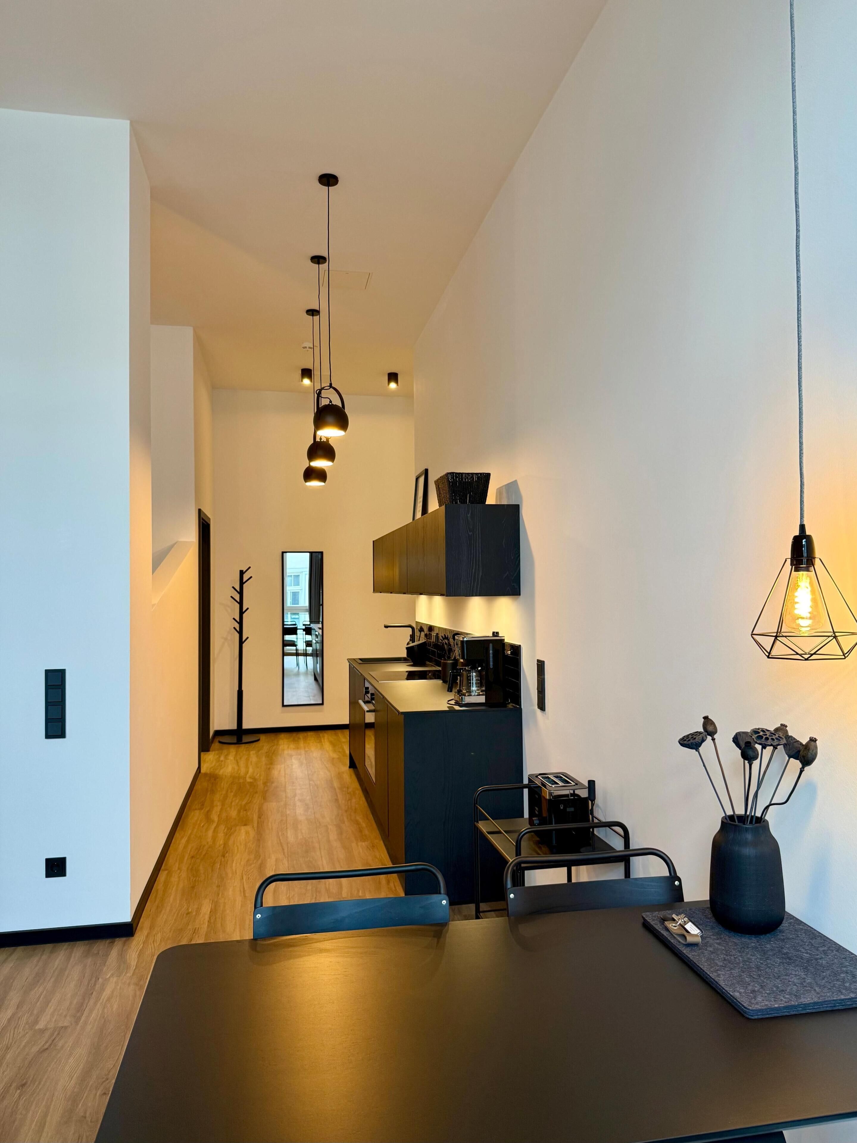 A modern kitchen with a dining area, featuring a dark countertop, hanging pendant lights, and a vase with dried flowers.