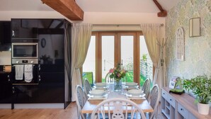 Un comedor con una mesa dispuesta para cuatro, una cocina con refrigerador y horno negros, y vistas al exterior a través de amplias ventanas.