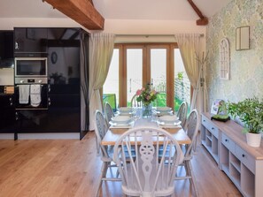 Ein Essbereich mit einem Tisch für vier Personen, eine Küche mit schwarzem Kühlschrank und Backofen sowie ein Blick ins Freie durch große Fenster.
