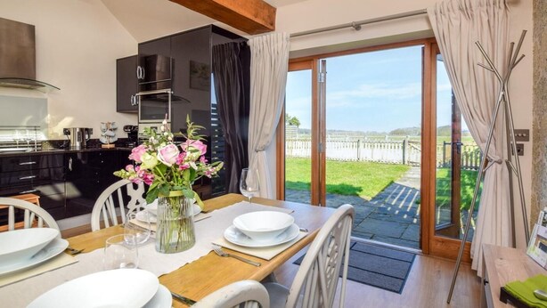 Un comedor con una mesa dispuesta para cuatro, una cocina con electrodomésticos modernos y vistas a un paisaje verde a través de una puerta de cristal.