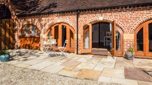 A brick building with arched doorways, a patio with stone tiles, and a table with chairs. - 1 Bed in Evesham (oc-89306) (Church Lench)