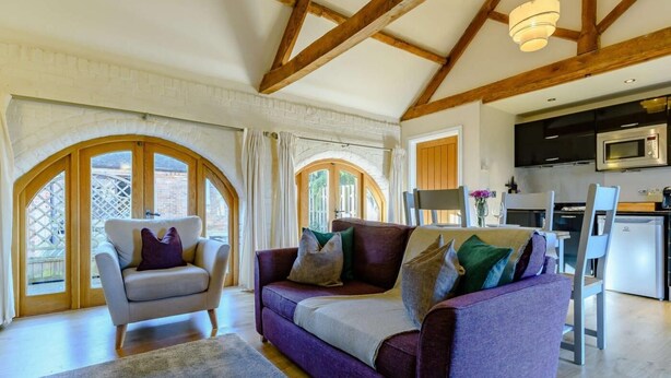 A spacious living area with a sofa, armchair, and dining table, wooden beams on the ceiling, and a kitchen with modern appliances. - 1 Bed in Evesham (oc-89306) (Church Lench)