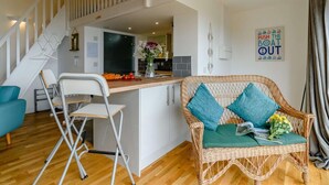 Un espacio habitable moderno con una isla central, un sofá de mimbre con cojines color turquesa, una silla plegable blanca y una escalera que conduce a un nivel superior.