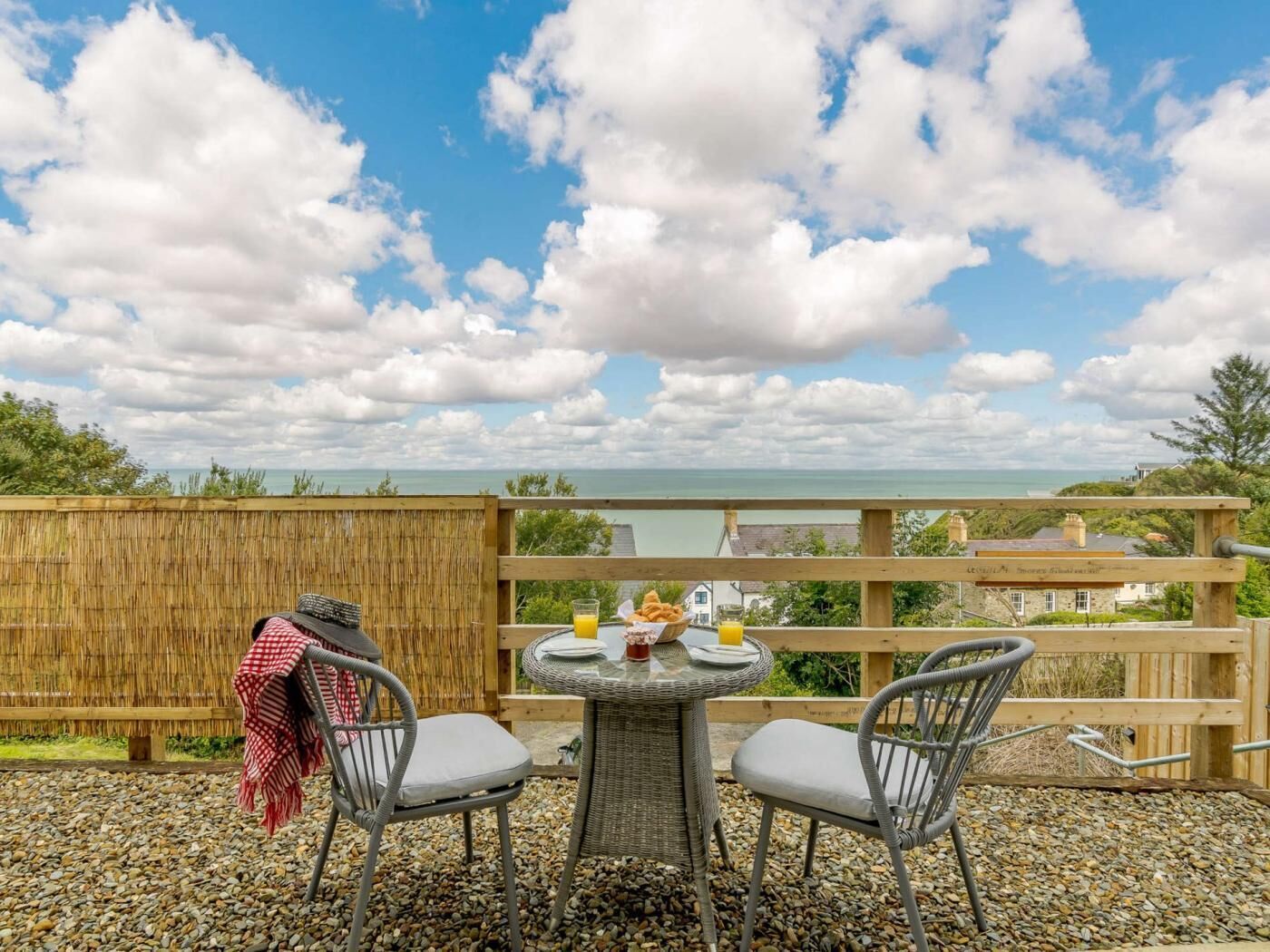 Een rieten tafeltje met twee stoelen, een rood-wit geruit kleed, en een glas sinaasappelsap op een balkon met uitzicht op het water en een verre kustlijn.