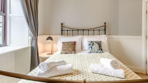 Cama bem arrumada com almofadas, abajur e janela com cortinas.