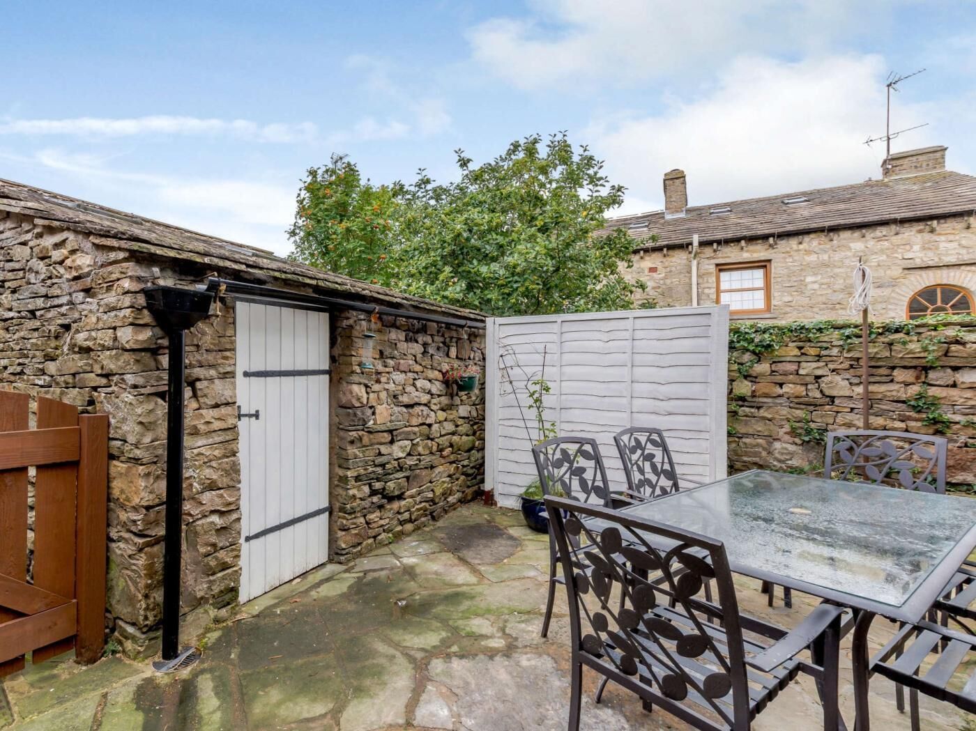 A stone building with a white door, a glass table with metal chairs, and a wooden bench.