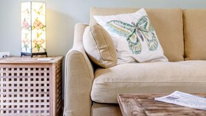 Ein beiges Sofa mit einem Kissen in Form eines Schmetterlings, ein Holztischchen mit einer Lampe und ein gerahmtes Bild an der Wand.