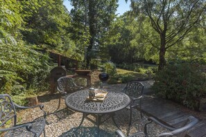 A patio with a round table and chairs, surrounded by greenery and a wooden deck. - 1 Bed in Newton Abbot (oc-bbell) (Hennock)