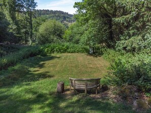 A grassy area with a wooden bench and a tree stump, surrounded by dense greenery and trees. - 1 Bed in Newton Abbot (oc-bbell) (Hennock)