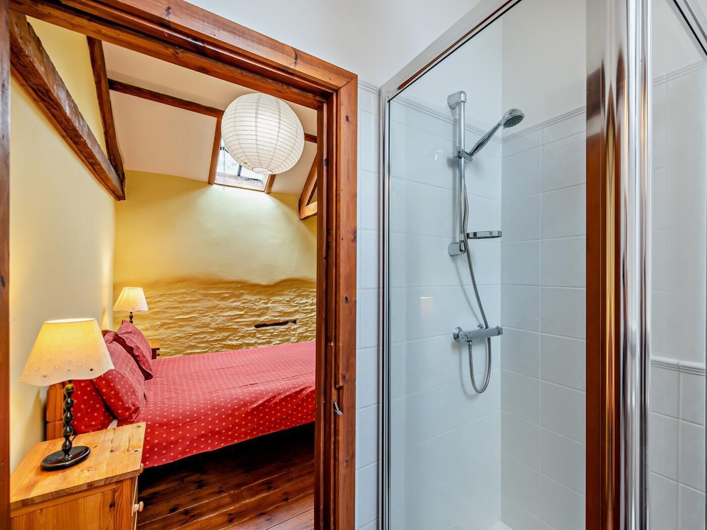 A bedroom with a wooden ceiling, a bed with a red bedspread, a nightstand with a lamp, and a glass shower enclosure.