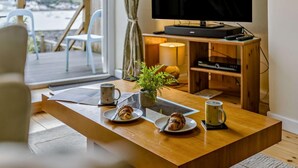 Una sala de estar moderna con una mesa de centro de madera, dos platos con pasteles, un televisor sobre un mueble de madera y vistas al exterior.