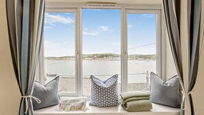 Una habitación con un amplio ventanal con vistas a una masa de agua, un radiador debajo y un asiento junto a la ventana con cojines.
