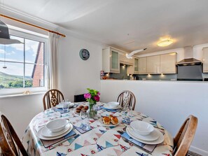 Een eethoek met een tafel gedekt voor vier personen, een raam met uitzicht en een keuken op de achtergrond.