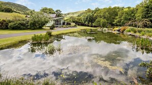 Une maison avec un étang devant, entourée de verdure et de collines.