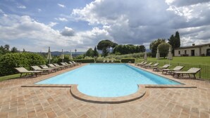 Uma piscina retangular com um deck ladrilhado, rodeada por espreguiçadeiras e guarda-sóis, sob um céu claro e com vegetação ao fundo.