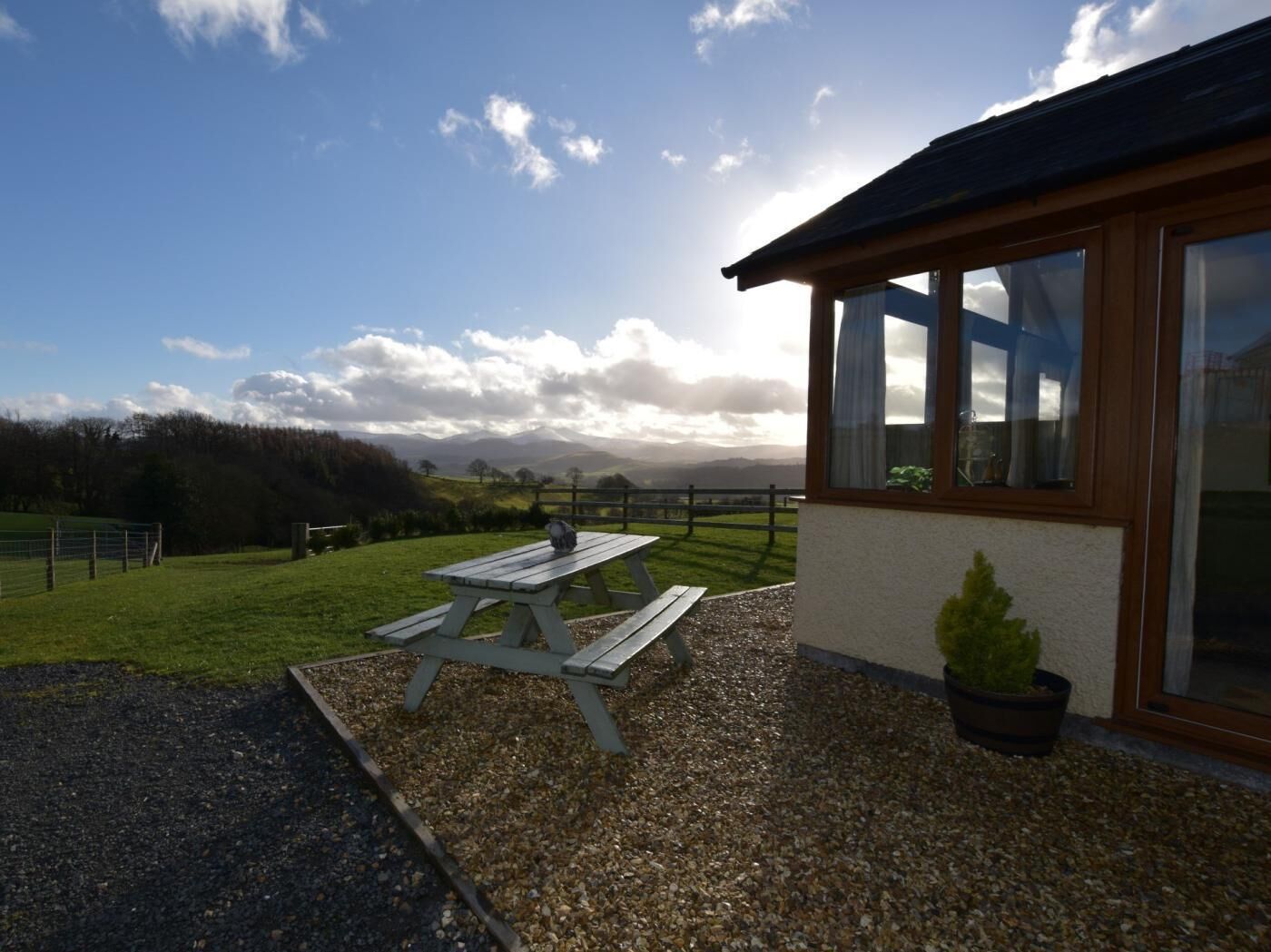 A building with a wooden exterior and large windows, a gravel patio with a picnic table, and a view of a green landscape with trees and hills.