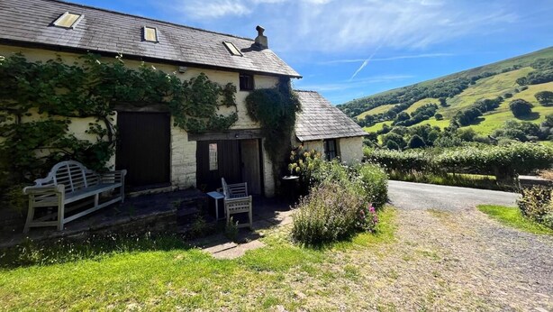 Ein Haus mit Schrägdach, einer Veranda mit Bank und Blick auf sanfte Hügel im Hintergrund.