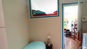 Ein kleines Zimmer mit einem Bett, einem Nachttisch, einer Lampe und Blick auf einen Essbereich durch eine offene Tür.