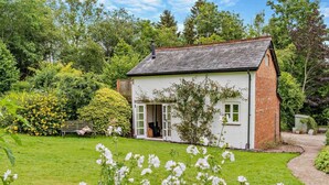 Ein kleines, eingeschossiges Haus mit weißer Fassade und einem Ziegelkamin, umgeben von einem gepflegten Garten mit blühenden Pflanzen und einer Rasenfläche.